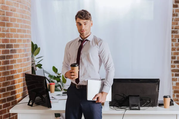 Giovane uomo d'affari che tiene caffè da asporto e laptop vicino ai computer in ufficio — Foto stock