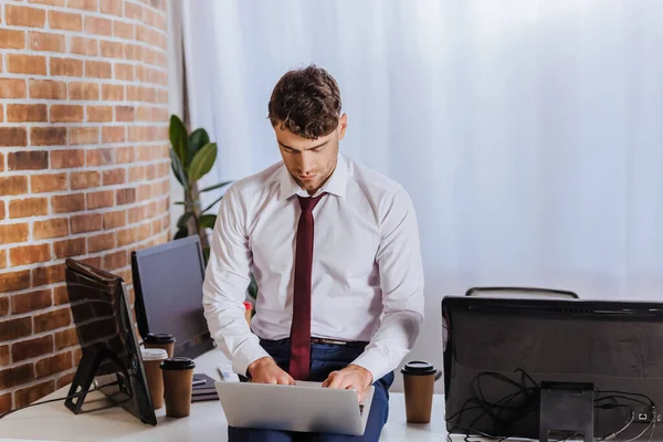 Empresário em desgaste formal usando laptop perto de café para ir e computadores no escritório — Fotografia de Stock