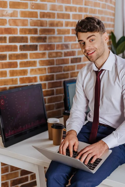 Smiling businessman using laptop near computer with charts and coffee to go on blurred background — Stock Photo
