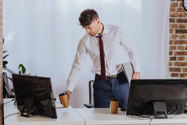 Молодой бизнесмен пьет кофе, держа ноутбук возле компьютеров в офисе — стоковое фото