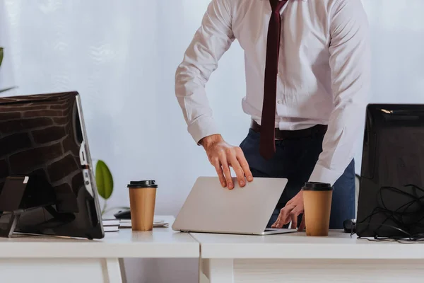 Vista recortada del hombre de negocios sosteniendo portátil cerca de café para llevar y computadoras en la oficina - foto de stock