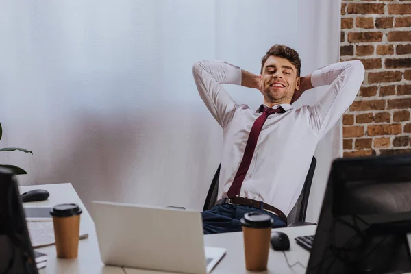 Hombre de negocios sonriente sentado cerca de computadoras, computadora portátil y café para ir en primer plano borroso - foto de stock