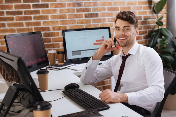 Uomo d'affari sorridente che parla su smartphone vicino a computer con grafici finanziari e caffè da asporto su sfondo sfocato — Foto stock