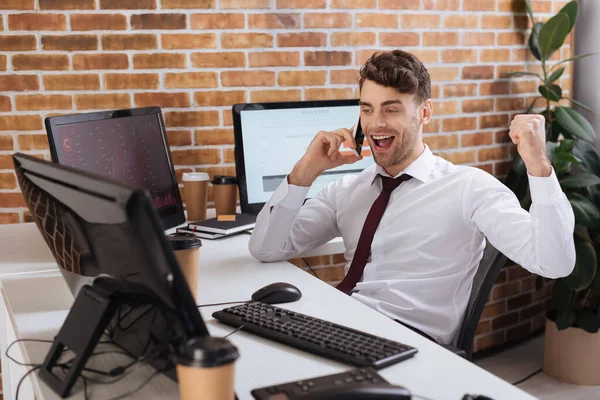 Allegro uomo d'affari che mostra sì gesto mentre parla su smartphone vicino al caffè per andare e computer in primo piano sfocato — Foto stock