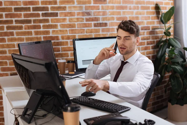 Businessman pointing at computer while talking on smartphone near coffee to go on blurred foreground — Stock Photo