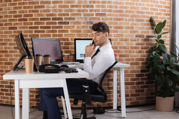 Hombre de negocios mirando a la cámara mientras habla en el teléfono inteligente cerca de café para llevar y computadoras en la mesa - foto de stock