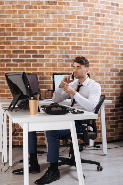 Young businessman pointing with finger at computer while talking on smartphone near takeaway coffee — Stock Photo