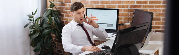 Amazed businessman talking on smartphone near computers and laptop, banner — Stock Photo