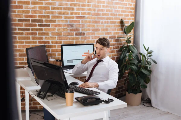 Hombre de negocios sorprendido hablando en el teléfono inteligente mientras revisa las acciones financieras en las computadoras - foto de stock