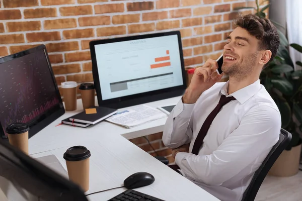 Lächelnder Geschäftsmann spricht auf Smartphone neben Computer mit Diagrammen von Finanzkursen im Büro — Stockfoto