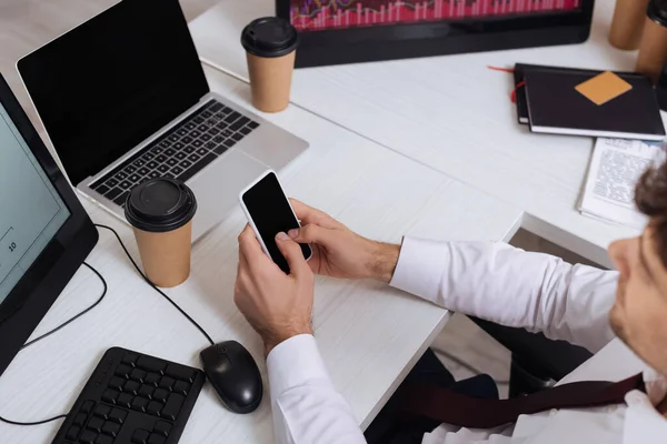 Visão de alto ângulo do smartphone com tela em branco em mãos de empresário em primeiro plano desfocado perto de computadores e café takeaway — Fotografia de Stock