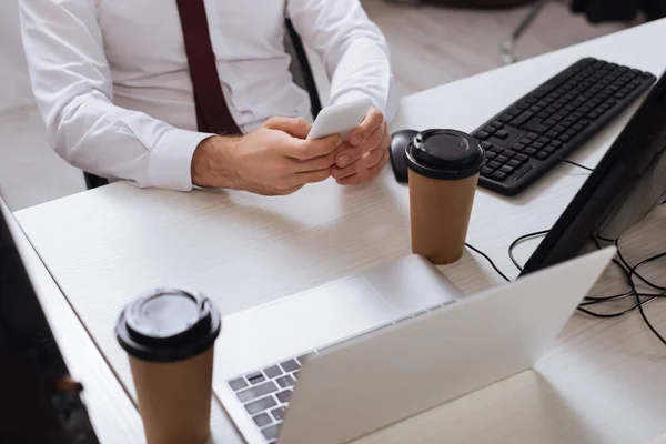 Vista recortada del hombre de negocios usando un teléfono inteligente cerca de computadoras y café para llevar en primer plano borroso - foto de stock