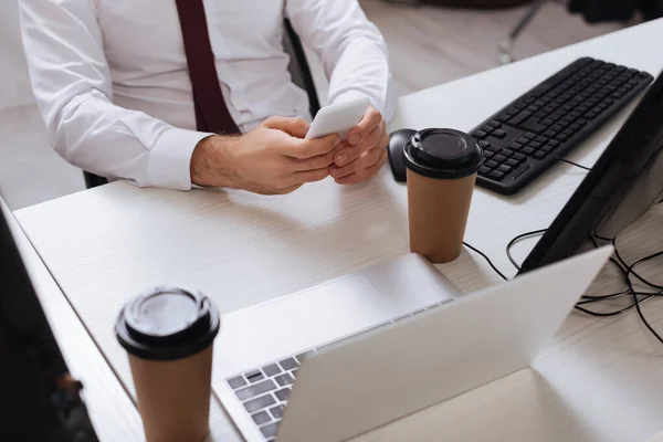 Vue recadrée de l'homme d'affaires à l'aide d'un smartphone près des ordinateurs et du café à emporter au premier plan flou — Photo de stock