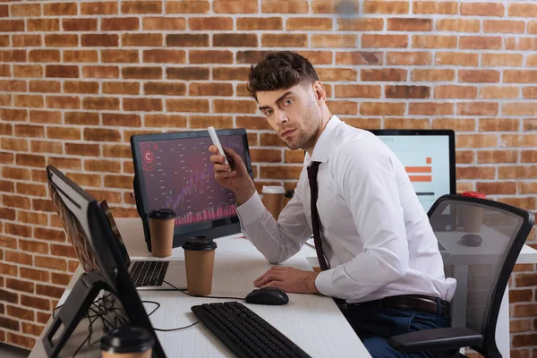 Homme d'affaires tenant smartphone près du café pour aller annoncer des ordinateurs avec des graphiques sur la table — Photo de stock