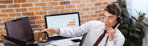 Cheerful businessman talking on telephone and pointing at computer while checking financial courses, banner — Stock Photo