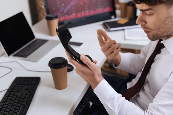 Empresario señalando con el dedo mientras sostiene auriculares telefónicos cerca de café para llevar y computadoras sobre fondo borroso - foto de stock