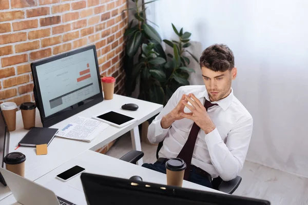 Joven hombre de negocios mirando el monitor de la computadora cerca de café para ir y gadgets en la oficina - foto de stock