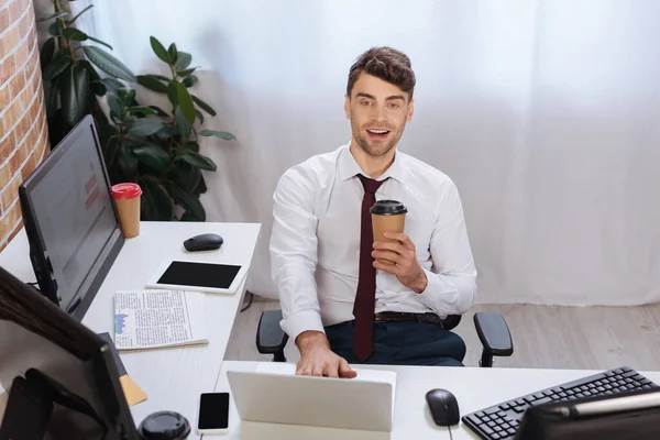 Un hombre de negocios sonriente sosteniendo café para acercarse a dispositivos y periódicos en la oficina - foto de stock