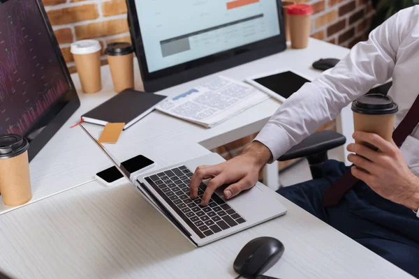 Ausgeschnittene Ansicht eines Geschäftsmannes, der im Büro Coffee to go hält und Finanzaktien am Laptop prüft — Stockfoto