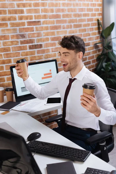 Empresário positivo com café takeaway sentado perto de computadores em primeiro plano desfocado — Fotografia de Stock