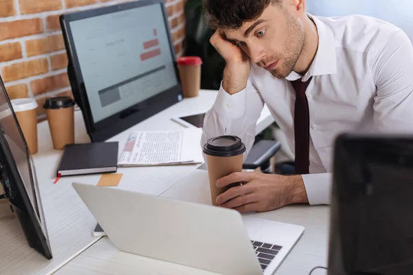 Tired businessman holding paper cup and checking financial stocks on laptop on blurred foreground — Stock Photo