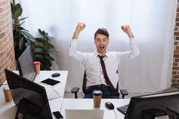 Emocionado hombre de negocios mostrando sí gesto cerca de las computadoras, café para llevar y periódico sobre fondo borroso - foto de stock