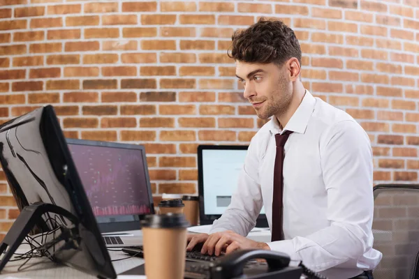 Businessman using computer near monitors with chars and coffee to go on blurred background — Stock Photo