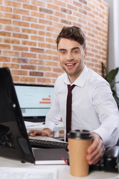 Homme d'affaires souriant prenant du café pour aller près de l'ordinateur sur le premier plan flou — Photo de stock