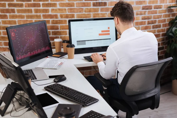 Vista posterior del hombre de negocios comprobando el mercado financiero cerca de la computadora con gráficos y periódico en la oficina - foto de stock