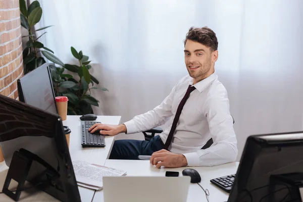 Uomo d'affari sorridente che guarda la telecamera vicino ai computer, il caffè da asporto e il giornale in primo piano sfocato — Foto stock