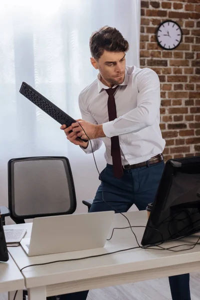 Homem de negócios irritado segurando teclado perto de computadores em primeiro plano desfocado no escritório — Fotografia de Stock
