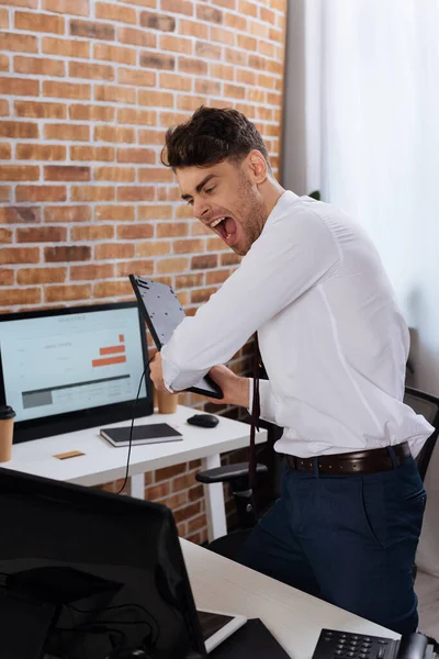 Homem de negócios louco segurando teclado e gritando perto do computador em primeiro plano desfocado — Fotografia de Stock