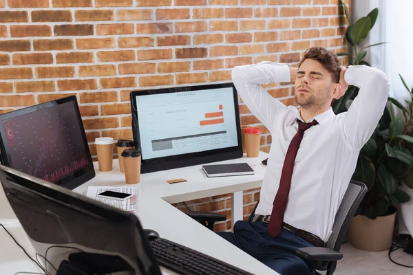 Hombre de negocios agotado sentado cerca de computadoras con cartas de cursos de finanzas en monitor - foto de stock