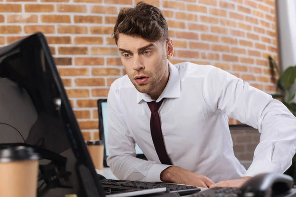 Businessman looking at computer monitor near telephone and coffee to go on blurred foreground — Stock Photo