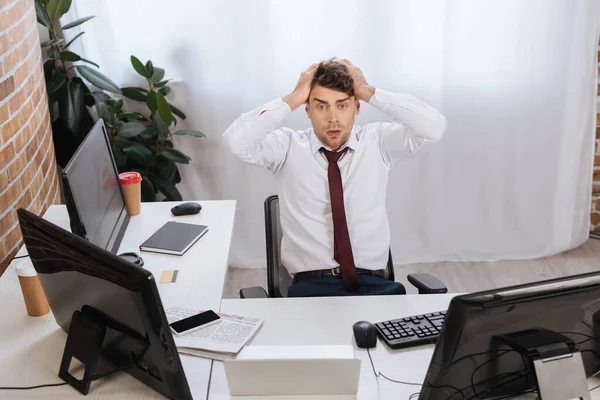Бизнесмен с руками возле головы смотрит в камеру рядом с устройствами и новостями на столе в офисе — стоковое фото