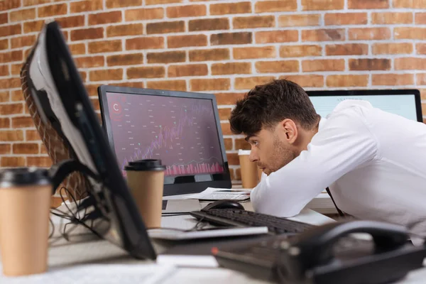 Concentrated businessman looking at computer with charts of finance stocks on monitor near telephone and takeaway coffee on blurred foreground — Stock Photo