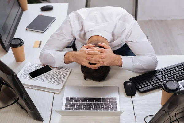 Overhead view of exhausted businessman sitting near computers, coffee to go and newspaper in office — Stock Photo