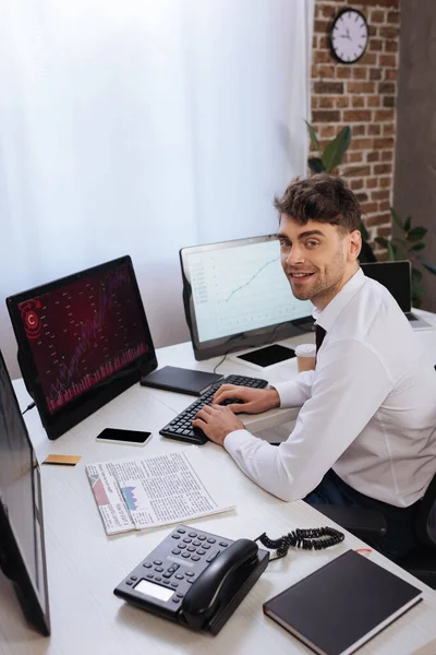 Uomo d'affari sorridente che utilizza il computer con grafici sul monitor vicino al telefono, giornali e notebook in primo piano sfocato — Foto stock