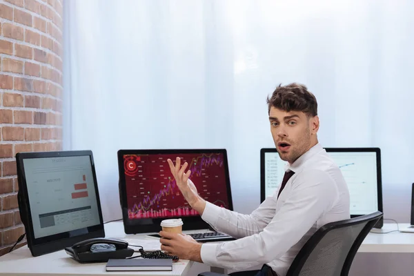 Erstaunter Geschäftsmann hält Kaffee zum Mitnehmen in der Nähe von Computern und Telefon auf dem Tisch — Stockfoto