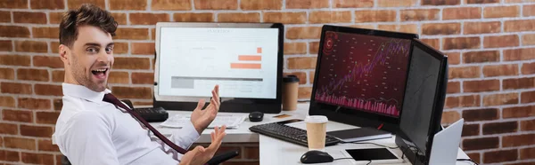 Empresário positivo olhando para a câmera perto de computadores e café para ir na mesa, banner — Fotografia de Stock