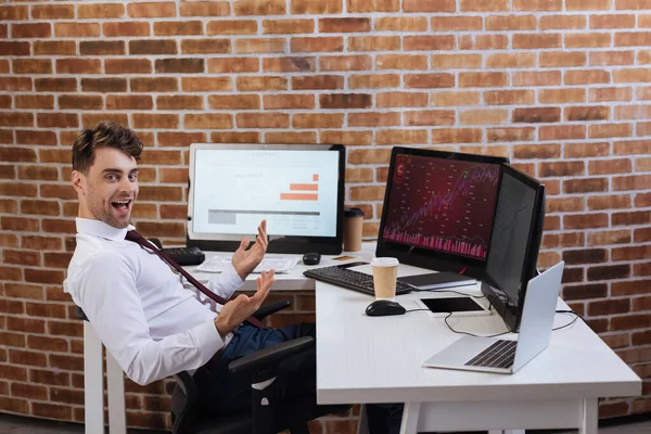 Cheerful businessman sitting near computers with graphs and takeaway coffee on table — Stock Photo