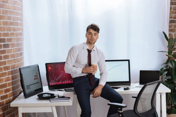 Businessman with coffee to go looking at camera near computers and telephone on table — Stock Photo