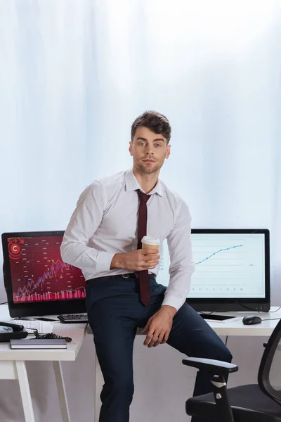 Young businessman holding coffee to go near computers with finance charts on blurred background — Stock Photo