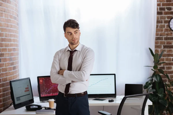 Uomo d'affari in piedi con le braccia incrociate vicino a computer su sfondo sfocato in ufficio — Foto stock