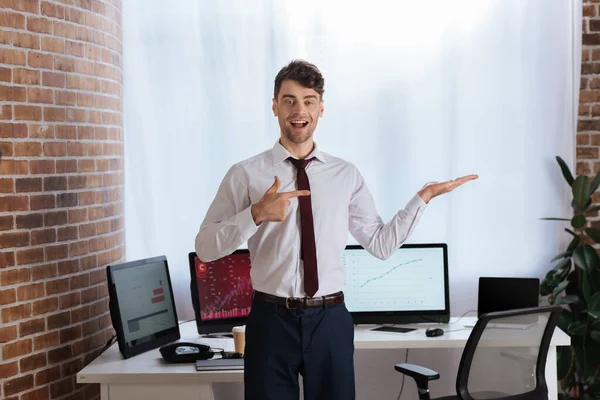 Empresário sorridente apontando com a mão e o dedo perto de computadores no fundo embaçado — Fotografia de Stock