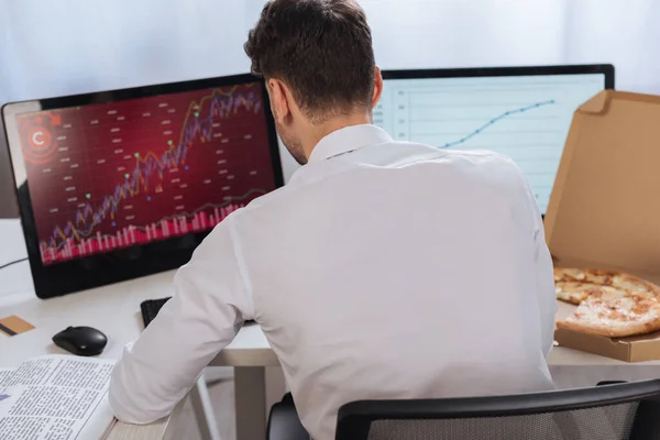 Vista trasera del hombre de negocios sentado cerca de computadoras con gráficos, periódico y pizza para llevar en un fondo borroso - foto de stock