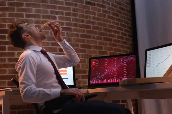 Бизнесмен ест пиццу на вынос рядом с компьютерами с графиками на размытом фоне в офисе вечером — стоковое фото