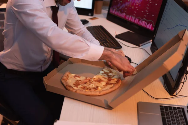 Vista ritagliata di uomo d'affari che prende gustosa pizza vicino laptop e computer su sfondo sfocato — Foto stock