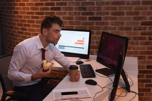 Homme d'affaires manger de la pizza avec du café à emporter tout en vérifiant les stocks sur les ordinateurs au bureau — Stock Photo