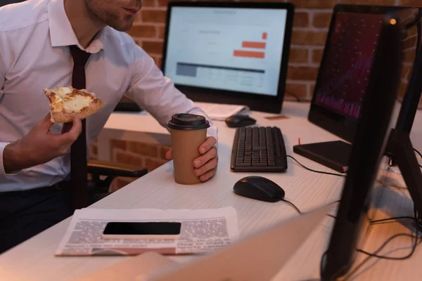 Vista recortada del hombre de negocios sosteniendo deliciosa pizza y café para ir cerca de computadoras, noticias y teléfonos inteligentes en la mesa - foto de stock